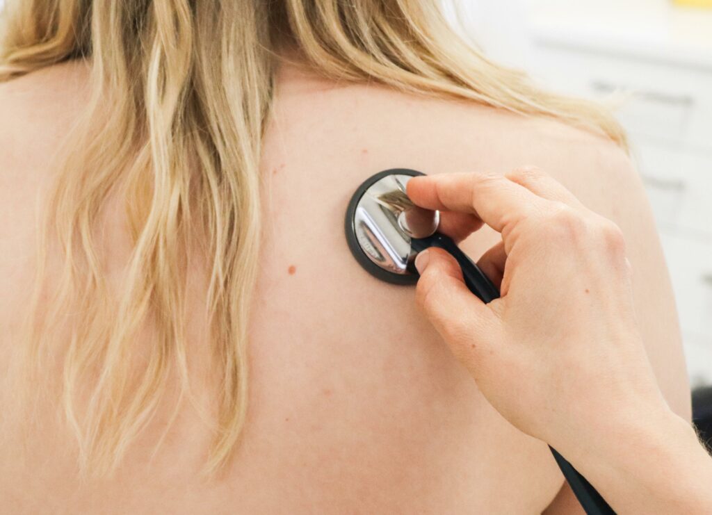 Rückansicht einer Patientin während einer pneumologischen Untersuchung im MVZ am Schlossplatz; sie sitzt im Behandlungsraum mit einem Stethoskop auf dem Rücken.